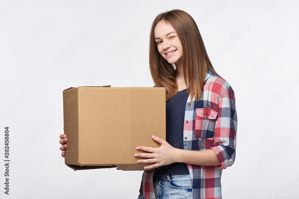 Delivery, relocation and unpacking. Happy playful young woman holding cardboard box winking at camera Delivery, relocation and unpacking. Happy playful young woman holding cardboard box winking at camera