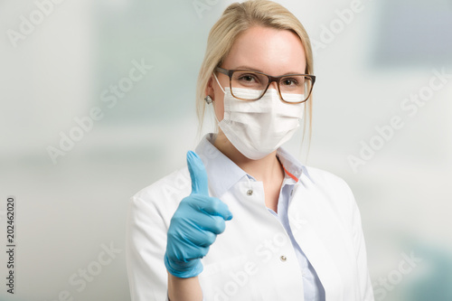 Junge hübsche Ärztin mit Mundschutz und hygienischen Handschuhen zeigt Daumen hoch photo