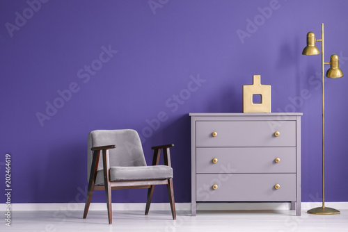 Ultra violet living room interior with retro armchair and chest of drawers next to a golden lamp. Real photo.