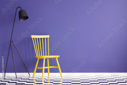 Bright, yellow chair standing next to a black lamp against purple wall in simple room interior. Real photo