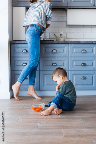 A cute little boy is sitting on the kitchen floor with his mother in the background