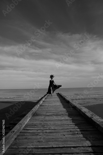 Beautiful woman in a dark luxury dress with big flower crown on head walking on a wooden pier. Dark clouds on background. Black and white