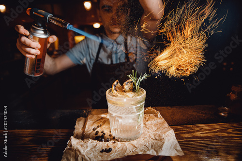 Barman prepares cocktail with orange and herbs in transparent glass on bar with alcohol. Uses burner with sparks. Dark background. photo