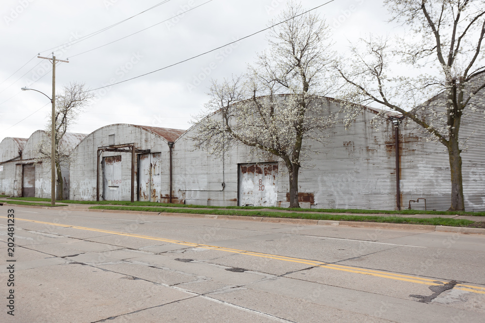 A blooming white crabapple trees next to a row of old warehouses. The warehouses are rusted and abandoned, with round roofs. A street runs beside them with a double yellow line and road repair patches