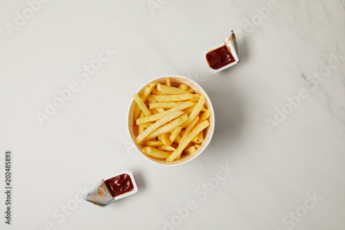 top view of french fries in bowl with containers of ketchup on white