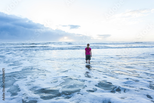 Young lady standing in ocean at the beach wearing shorts and t-shirt, with blonde hair. It is sunrise, and the sun is beginning to come over the horizon.