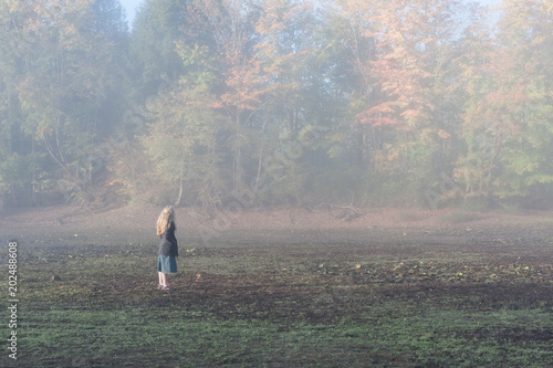 Fototapeta Naklejka Na Ścianę i Meble -  A girl with blonde hair standing in field on a foggy morning, with autumn leaves and forest in background. The woman is wearing a denim skirt and a jacket.