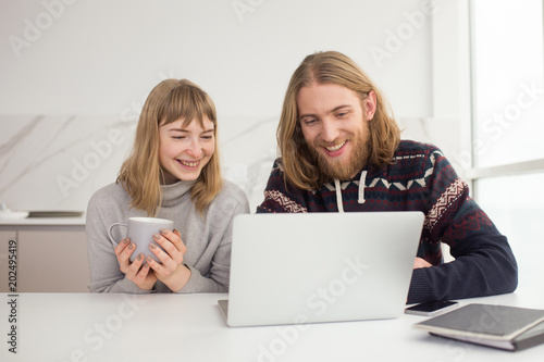 Portrait of young joyful couple sitting together with mug and laptop at home isolated