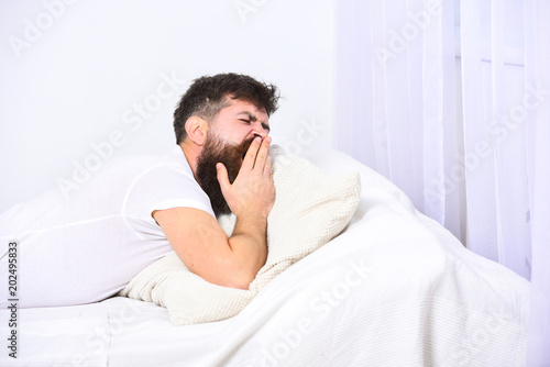 Man in shirt yawning while laying on bed, white wall and curtain on background. Sleepyhead concept. Guy on sleepy tired face yawning. Macho with beard and mustache yawning, relaxing, having nap, rest.