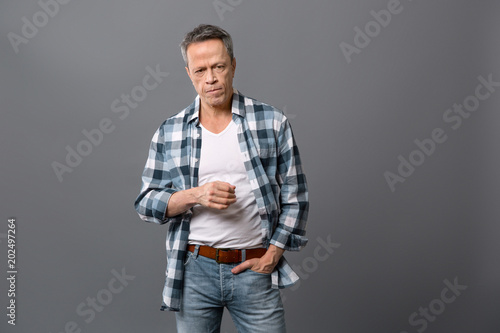 Brutal man. Serious aged man putting hand in his pocket while standing against grey background
