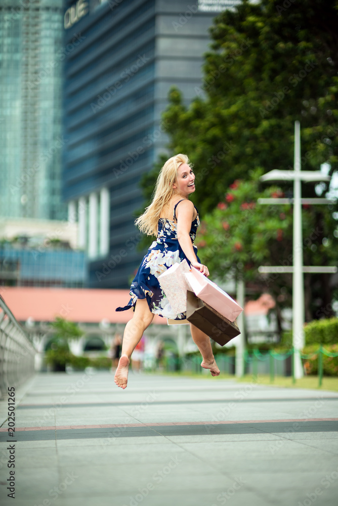 The Joy of Shopping... Excited Beautiful Woman on air, wearing casual blue dress with flowers, shopping bags in the streets with skyscrapers