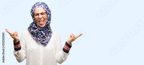 Middle age muslim arab woman wearing hijab looking at camera showing tong and making victory sign with fingers isolated blue background