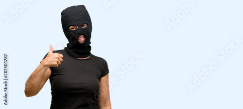 Burglar terrorist woman wearing balaclava ski mask smiling broadly showing thumbs up gesture to camera, expression of like and approval isolated blue background