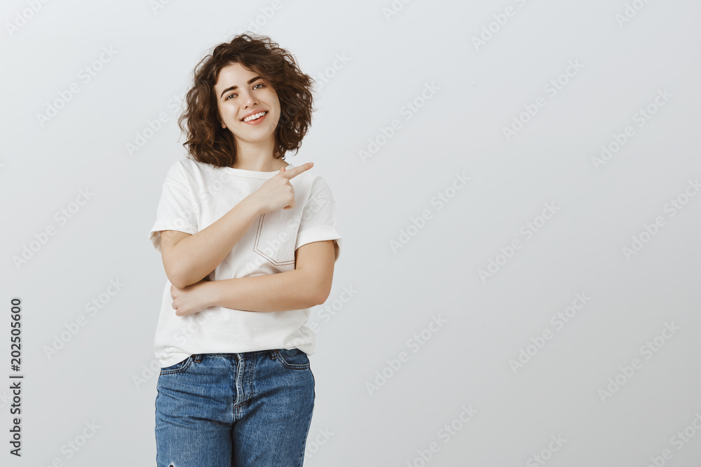 Friend suggesting to visit specialist she knows. Portrait of charming friendly caucasian female with short curly hair, pointing at upper right corner and smiling with assured expression Friend suggesting to visit specialist she knows. Portrait of charming friendly caucasian female with short curly hair, pointing at upper right corner and smiling with assured expression