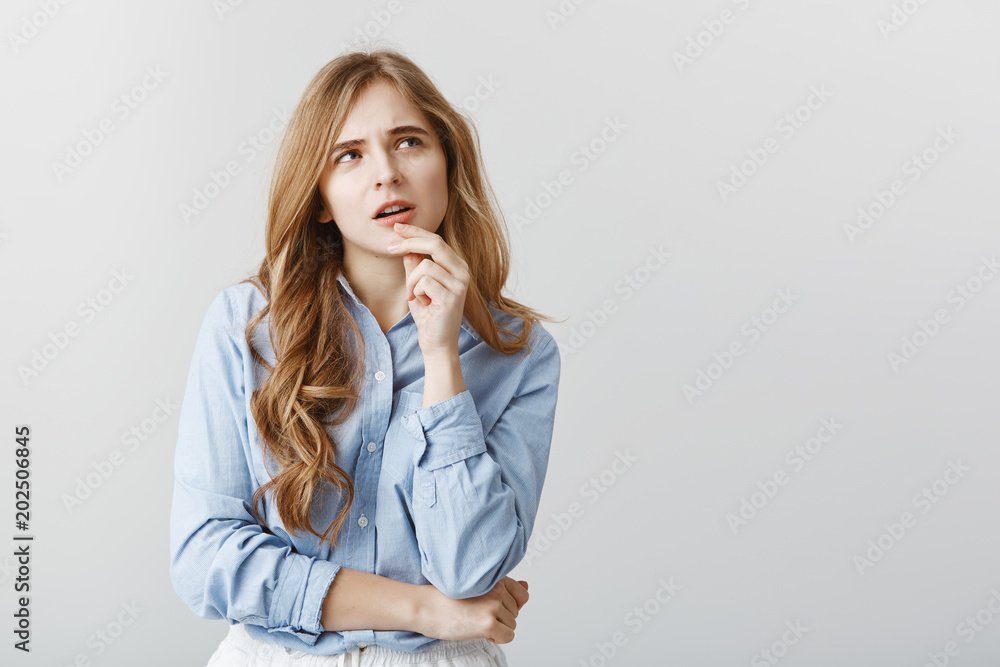 Continue diet or order pizza. Portrait of questioned thinking attractive woman with blond hair and curls, touching lip and looking up with thoughtful focused expression, having idea over gray wall Continue diet or order pizza. Portrait of questioned thinking attractive woman with blond hair and curls, touching lip and looking up with thoughtful focused expression, having idea over gray wall