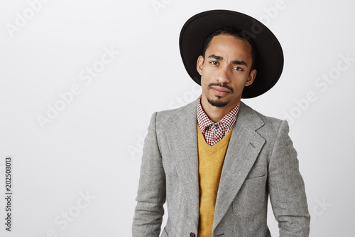 Businessman on meeting with partners. Serious attractive successful dark-skinned employer in trendy hat and formal outfit, focusing on conversation, talking about deal between companies over gray wall