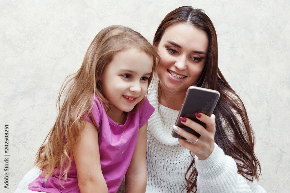 Young woman and girl are looking together at the mobile phone screen. They are sitting at a table on which there is a laptop. It's mom and daughter. Young woman and girl are looking together at the mobile phone screen. They are sitting at a table on which there is a laptop. It's mom and daughter.
