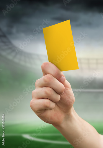 Hand holding up yellow card against football pitch in large stadium 