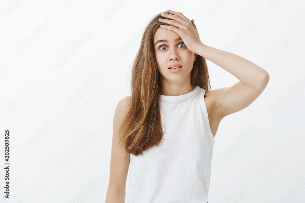 Confused questioned attractive woman with fair hair, staring cluelessly at camera and touching forehead with palm, remembering something or recalling what she forgot over gray wall, feeling anxious