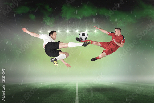 Football players tackling for the ball against football pitch under green sky