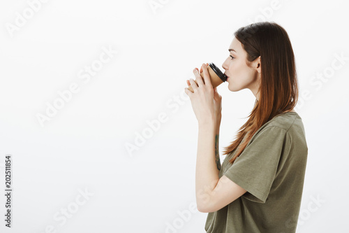 Attractive busy woman cannot start day without cup of coffee. Profile portrait of pretty adult brunette in casual t-shirt, drinking tea and enjoying cozy evening, looking outside cafe window on street