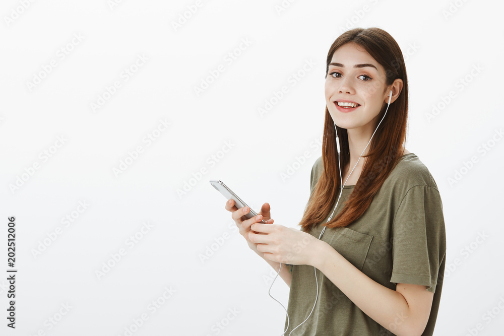 Studio portrait of pleased confident young european woman in dark-green t-shirt, gazing at camera while picking song in smartphone, listening music in earphones over gray background Studio portrait of pleased confident young european woman in dark-green t-shirt, gazing at camera while picking song in smartphone, listening music in earphones over gray background