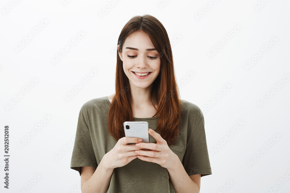 Happy loving girlfriend texting with satisfied expression. Portrait of pleased playful young girl in casual outfit, holding smartphone and smiling broadly, gazing at screen with satisfaction Happy loving girlfriend texting with satisfied expression. Portrait of pleased playful young girl in casual outfit, holding smartphone and smiling broadly, gazing at screen with satisfaction
