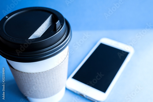 white paper kraft disposable cup for coffee with black plastic lid and white mobile phone on blue background. photo