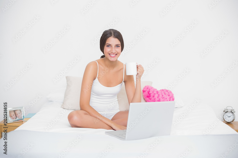 Cheerful young brown haired model in white pajamas drinking coffee while using laptop Cheerful young brown haired model in white pajamas drinking coffee while using laptop