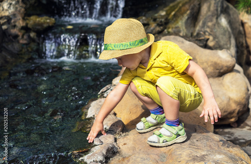   hild kid boy 3 year in straw hat and a yellow T-shirt  touches his hand to the water in the pond. Vacations and Walking in zoo