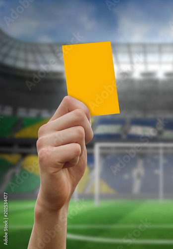 Hand holding up yellow card against football pitch in large stadium