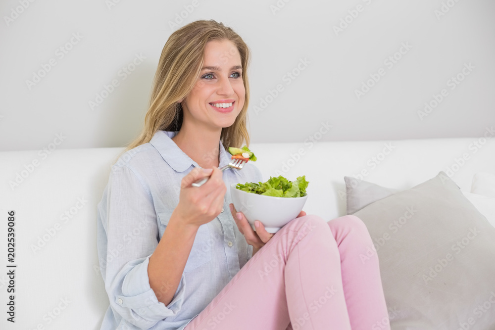 Happy casual blonde sitting on couch holding salad bowl Happy casual blonde sitting on couch holding salad bowl