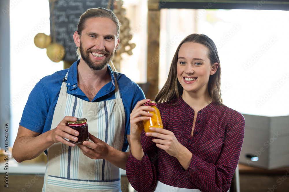 Shop assistants interacting while holding jam and pickle jar at grocery shop Shop assistants interacting while holding jam and pickle jar at grocery shop
