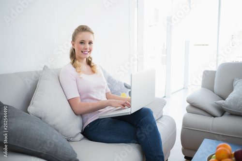 Casual smiling blonde sitting on couch using laptop