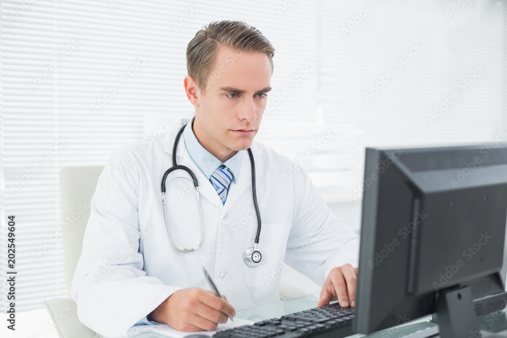 Doctor writing a note while using computer at medical office Doctor writing a note while using computer at medical office