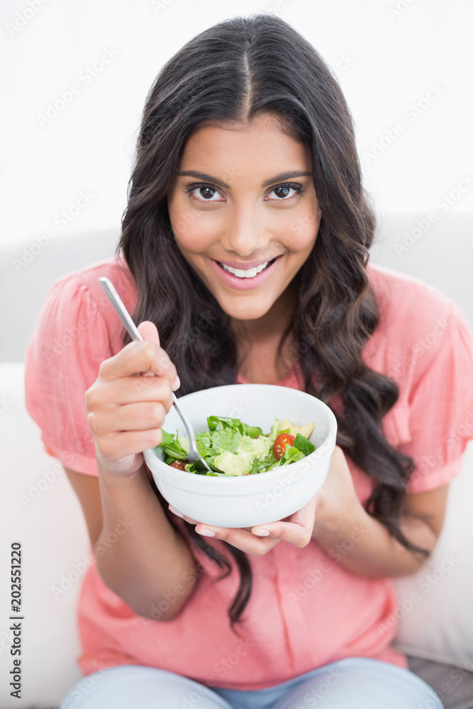 Cheerful cute brunette sitting on couch holding salad bowl Cheerful cute brunette sitting on couch holding salad bowl