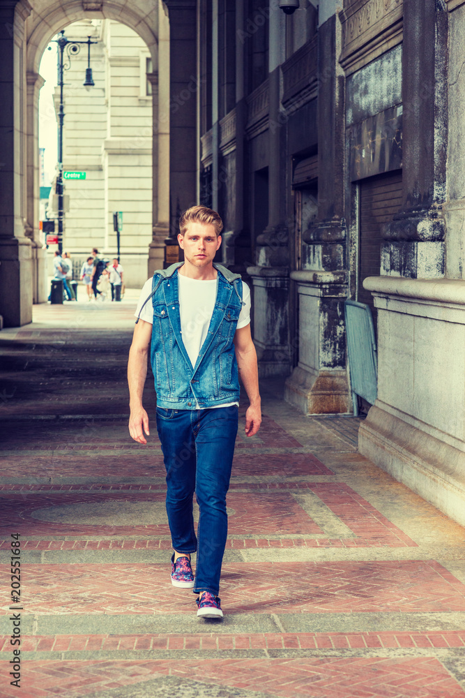 Young American Man with little beard traveling in New York, wearing blue Denim hoody sleeveless vest jacket, white T shirt, jeans, casual shoes, walking through small, narrow vintage street. Young American Man with little beard traveling in New York, wearing blue Denim hoody sleeveless vest jacket, white T shirt, jeans, casual shoes, walking through small, narrow vintage street.