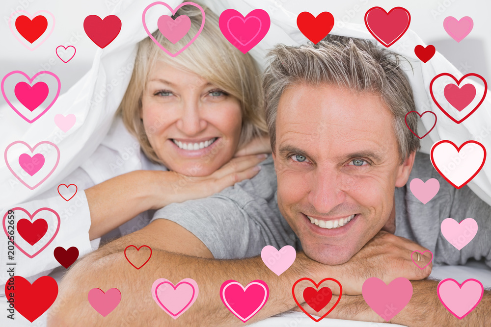 Couple smiling under the covers against valentines heart design Couple smiling under the covers against valentines heart design