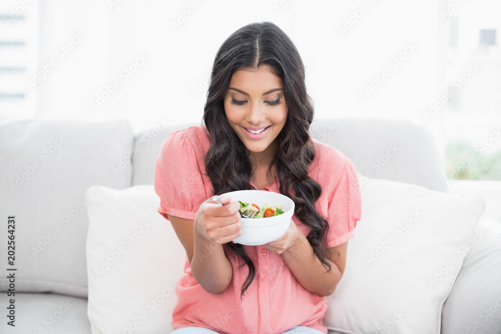 Content cute brunette sitting on couch holding salad bowl Content cute brunette sitting on couch holding salad bowl
