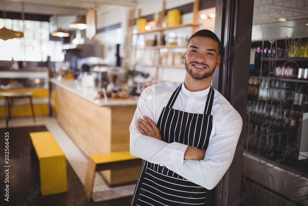 Portrait of smiling young handsome waiter with arms crossed leaning on cabinet