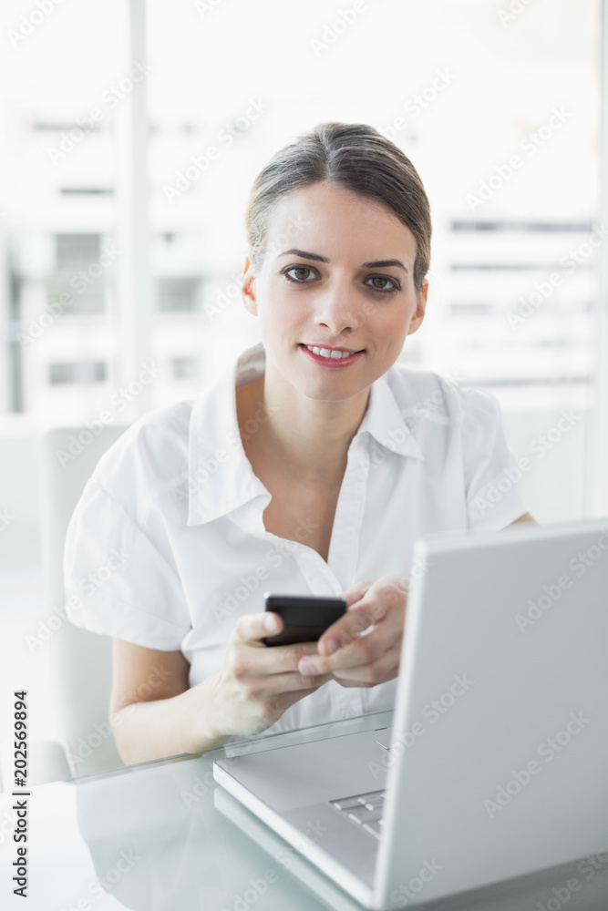 Lovely businesswoman holding her smartphone smiling softly at camera Lovely businesswoman holding her smartphone smiling softly at camera