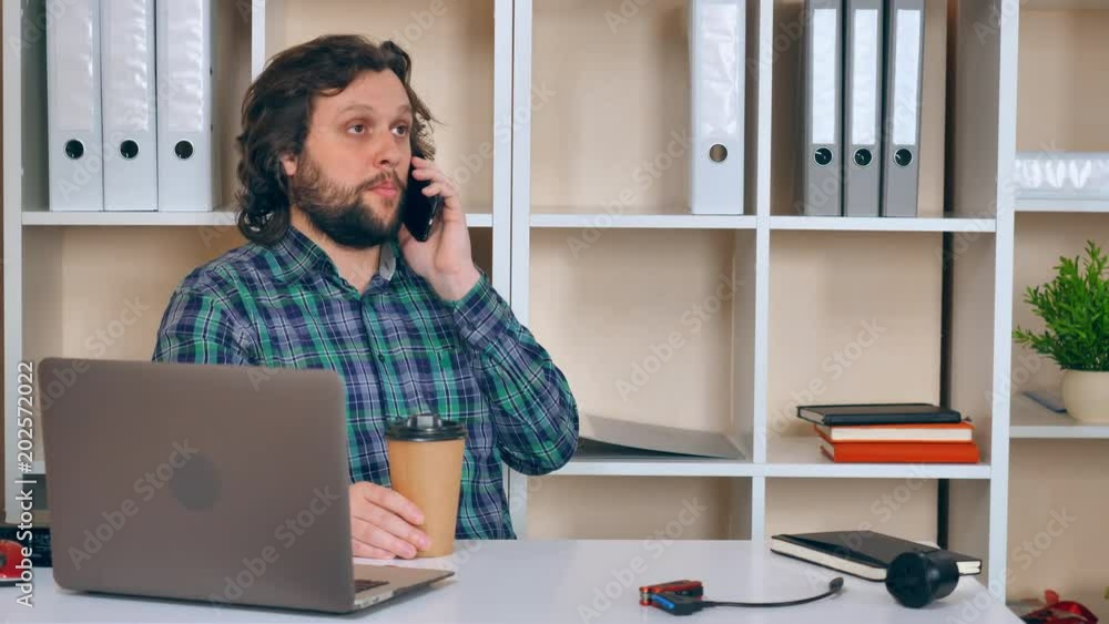 hipster man has phone conversation in casual small office. handsome guy with beard using smartphone drinking coffee sitting at the white desk in workplace hipster man has phone conversation in casual small office. handsome guy with beard using smartphone drinking coffee sitting at the white desk in workplace