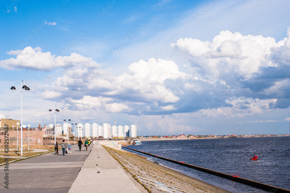 People walk along the promenade on the background of a large river and beautiful clouds