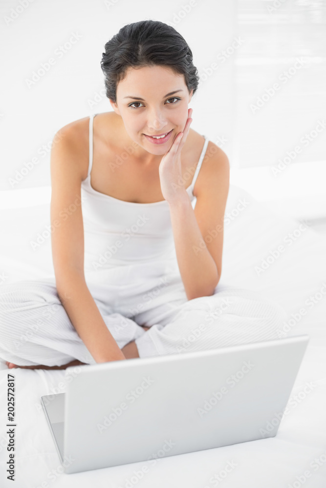 Cheerful casual brunette in white pajamas sitting on her bed and using a laptop Cheerful casual brunette in white pajamas sitting on her bed and using a laptop