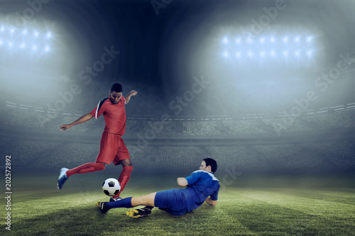 Football players tackling for the ball against large football stadium under blue sky © vectorfusionart
