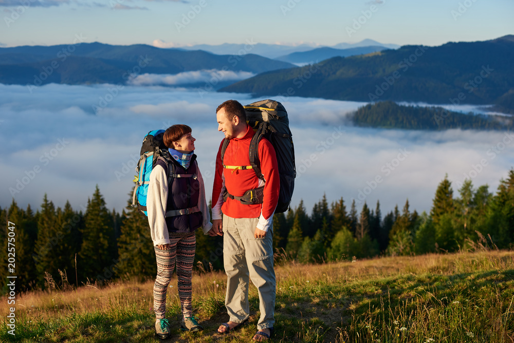 A man and a woman with backpacks look at each other in the rays of a sunset against the backdrop of the beautiful scenery of the forest, mountains and mist lying on them