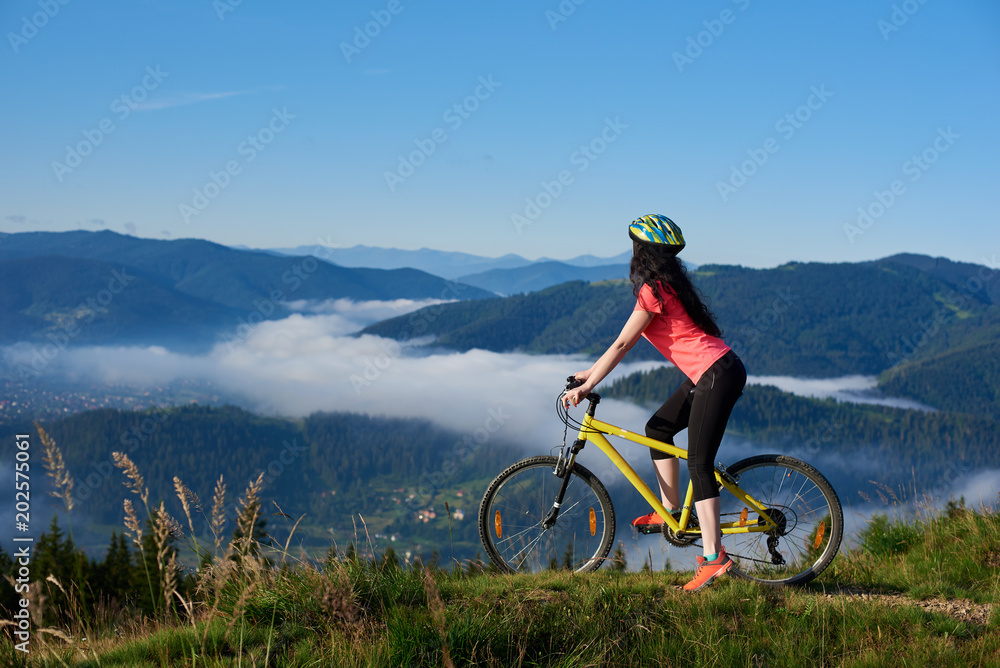 Sporty female cyclist riding on yellow bicycle on the top of mountain, wearing helmet and red red t-shirt, enjoying morning haze in valley, forests on the blurred background. Outdoor sport activity