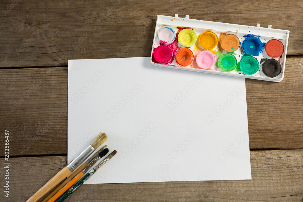 Colorful palette paintbrushes and blank paper on wooden surface Colorful palette paintbrushes and blank paper on wooden surface