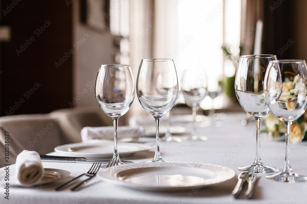 Table setting in the restaurant, glasses in the foreground. Luxury wedding reception. Flower arrangement on table in restaurant. Stylish decor and adorning. Table setting in the restaurant, glasses in the foreground. Luxury wedding reception. Flower arrangement on table in restaurant. Stylish decor and adorning.