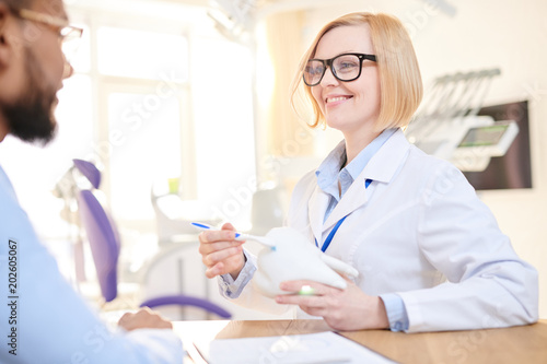 Friendly blond-haired dentist wearing white coat and eyeglasses sitting opposite her male patient and explaining him stages of treatment with help of tooth model